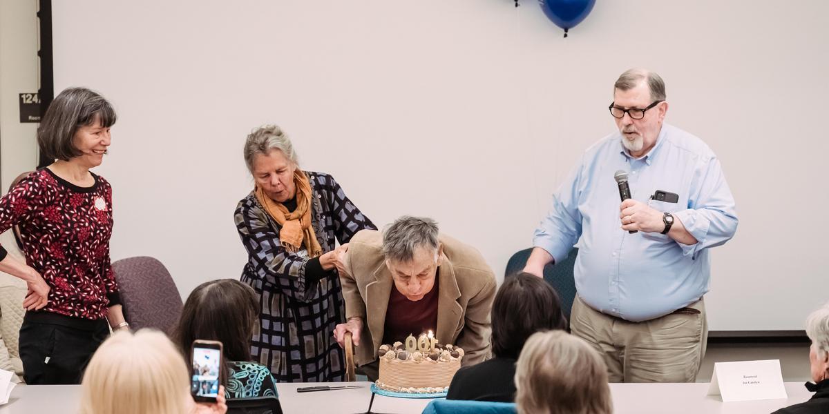 Mel bending over to blow out the candles on a 100th birthday cake as friends look on.