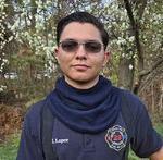 Jeydan Lopez, wearing a t-shirt from his fire department and holding a yellow helmet. He is standing in front of a cherry blossom tree.