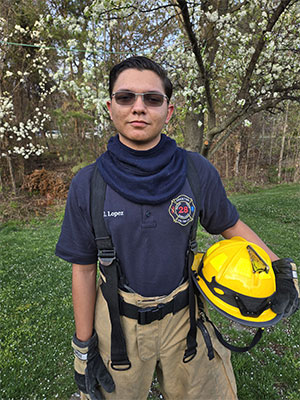 Jeydan Lopez, wearing a t-shirt from his fire department and holding a yellow helmet. He is standing in front of a cherry blossom tree.