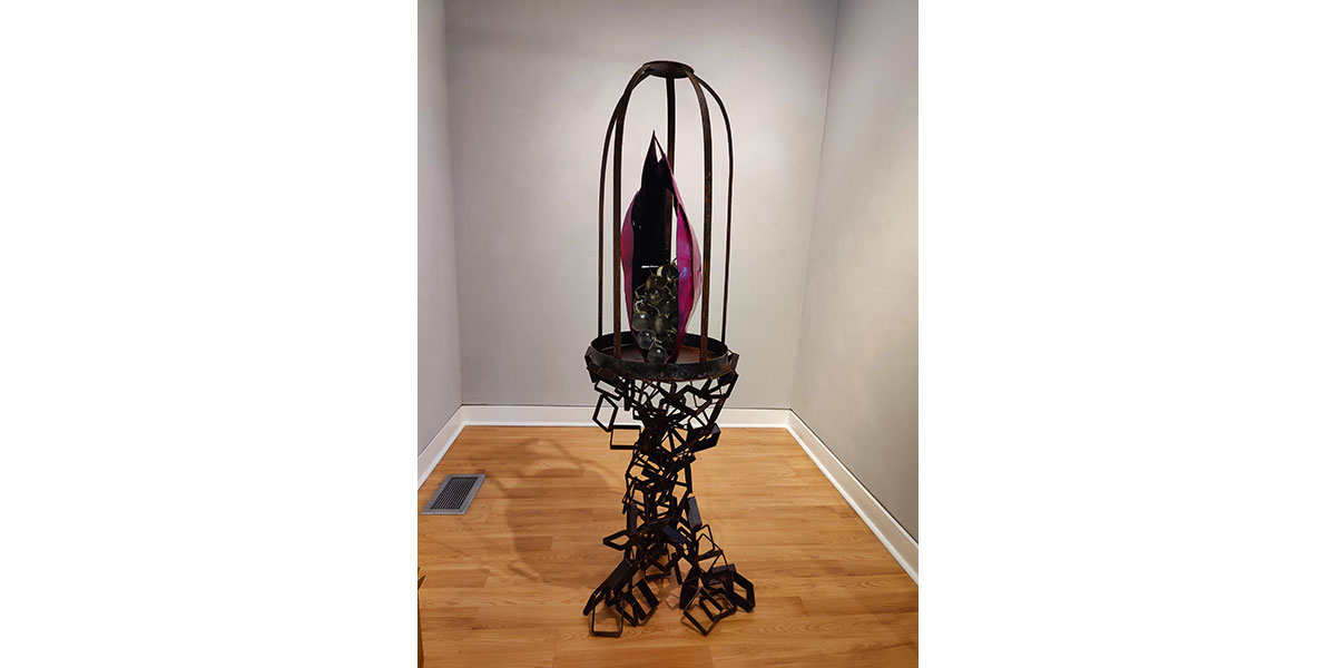 A metal sculpture displayed in a gallery corner features an abstract, geometric base supporting a circular platform with tall, curved metal rods forming a cage-like structure around a central purple and black form.