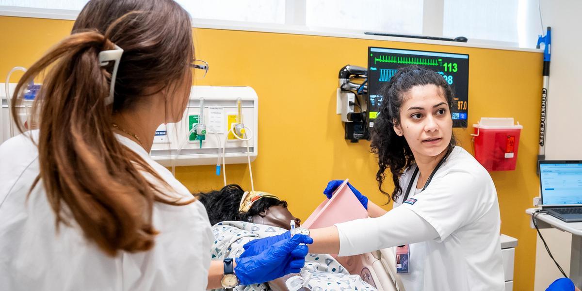 Two nursing students practice patient care in a clinical simulation room. One student wears gloves and holds a medical device while assisting a patient mannequin in a hospital bed, with medical monitors and equipment visible on the wall behind them.