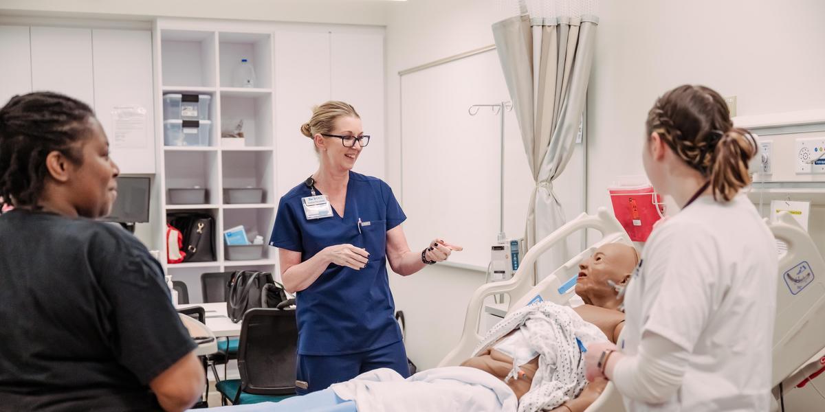 In a medical simulation lab, an instructor in blue scrubs guides two students around a medical mannequin lying in a hospital bed.