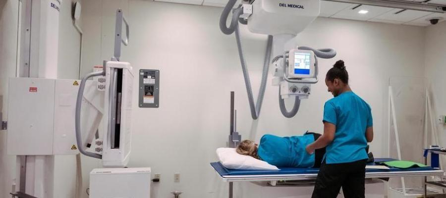 In a medical imaging lab, a student in blue scrubs assists a person lying on an examination table. They are positioned beneath a large, ceiling-mounted X-ray machine.