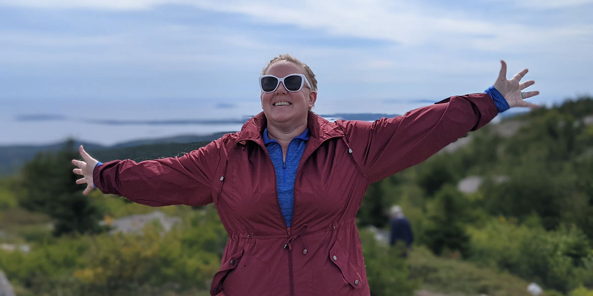 Sage Perkins with their arms outstretch with greenery and watery behind them